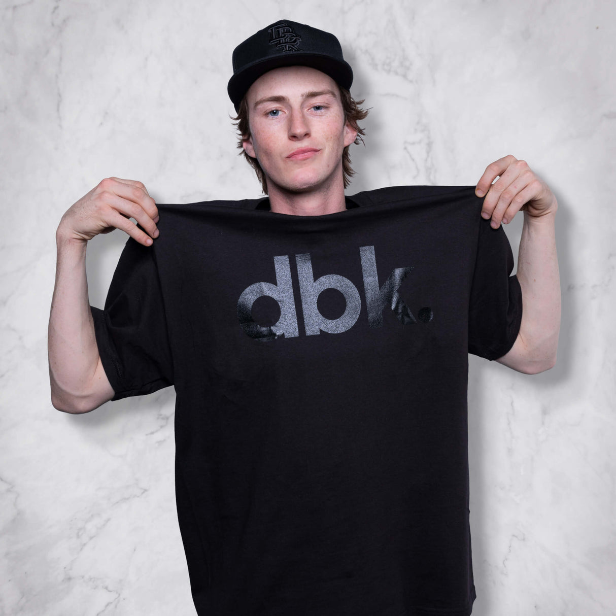 Black dbk graphic T-shirt held up by man wearing a cap