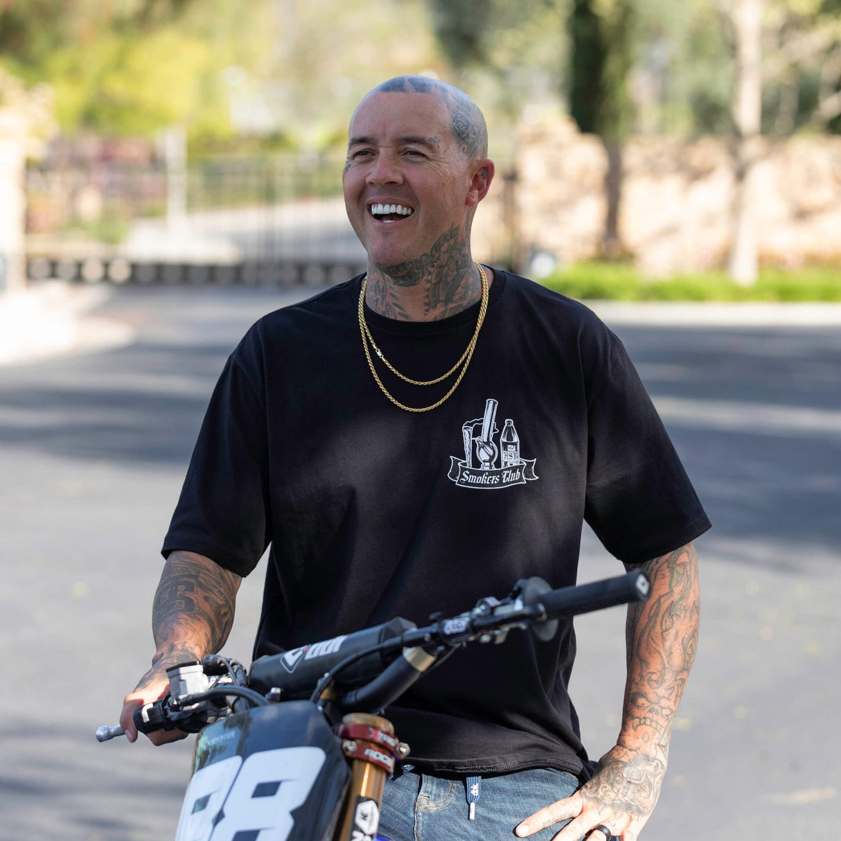 Black graphic T-shirt with Smokes Club bottle logo on the chest worn by a rider