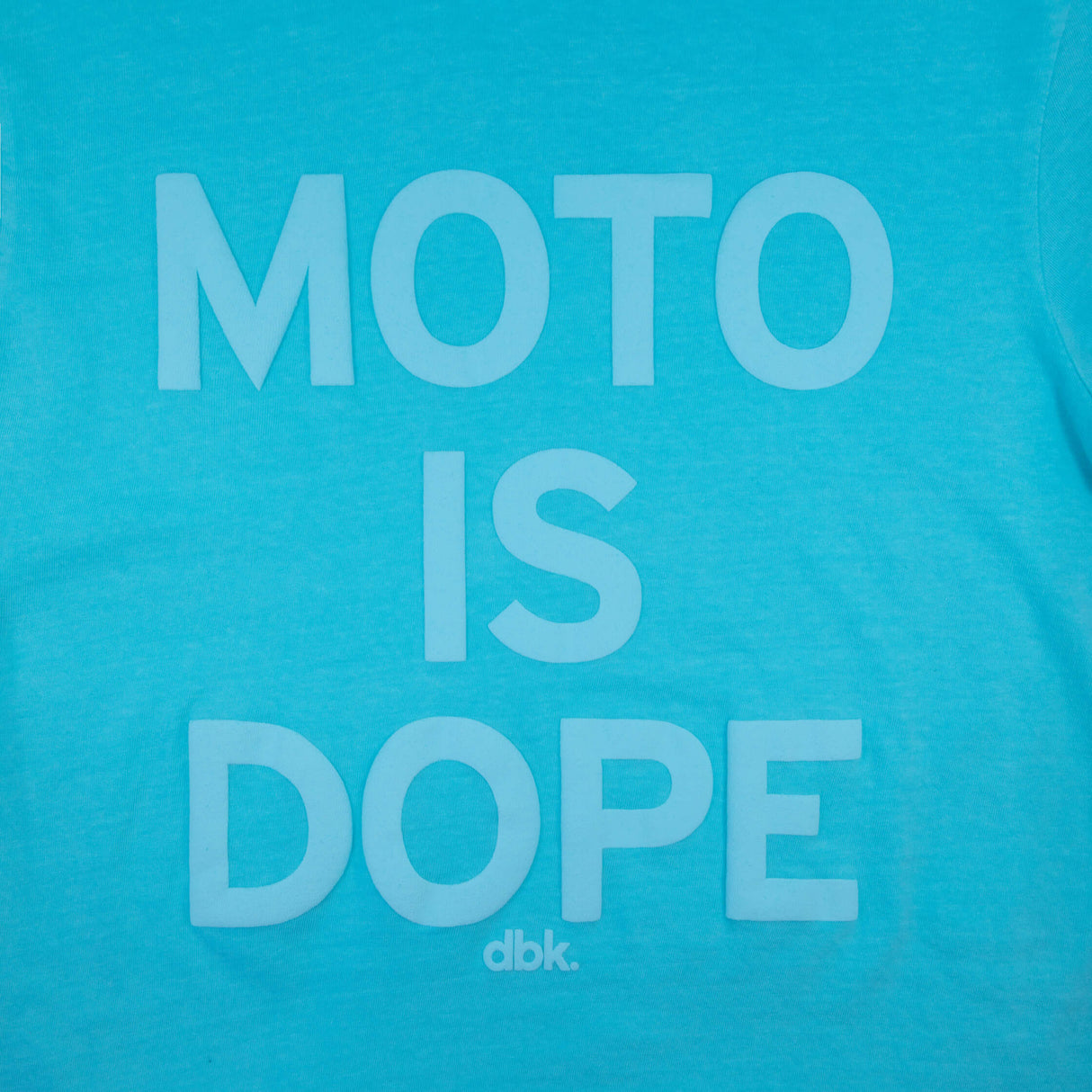Women’s graphic T-shirt in teal with centered white “Moto is Dope” text print and dbk. mark.