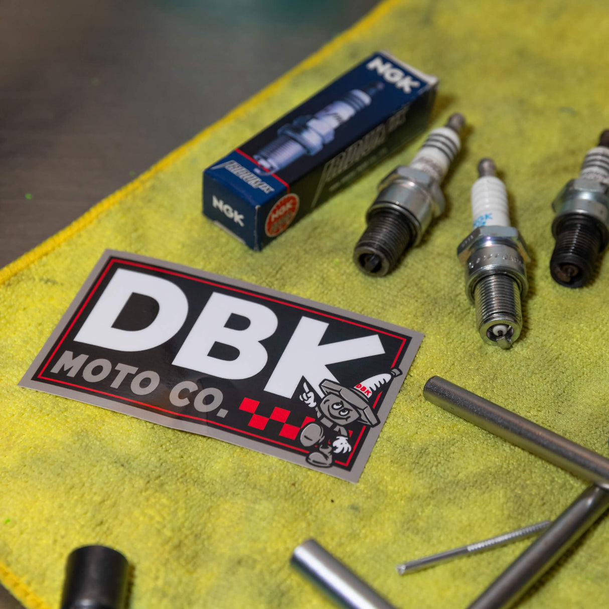 DBK Moto Co sticker with DBK letters and cartoon character, lying on yellow cloth