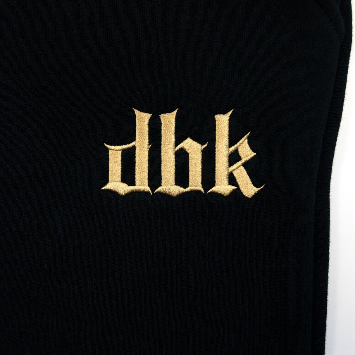 Black sweatpants leg with gold embroidered dbk logo centered on the fabric.