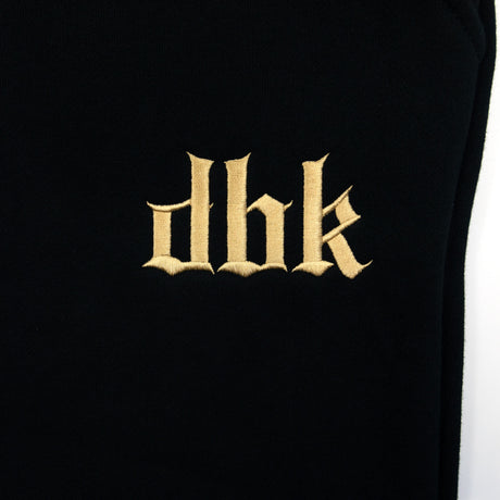 Black sweatpants leg with gold embroidered dbk logo centered on the fabric.