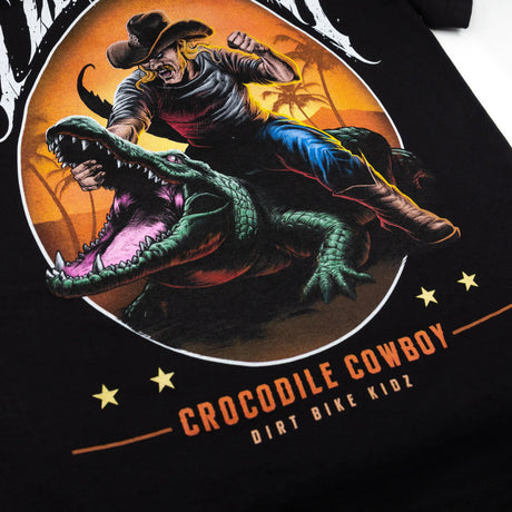 Black youth T-shirt close-up of circular cowboy-and-crocodile art with “CROCODILE COWBOY” below.