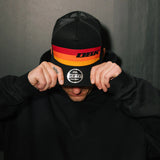 Black trucker cap with orange-red-yellow stripe and DBK logo, head-on view