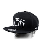 Black baseball cap with white 'DBK' logo and small white X graphic on the side
