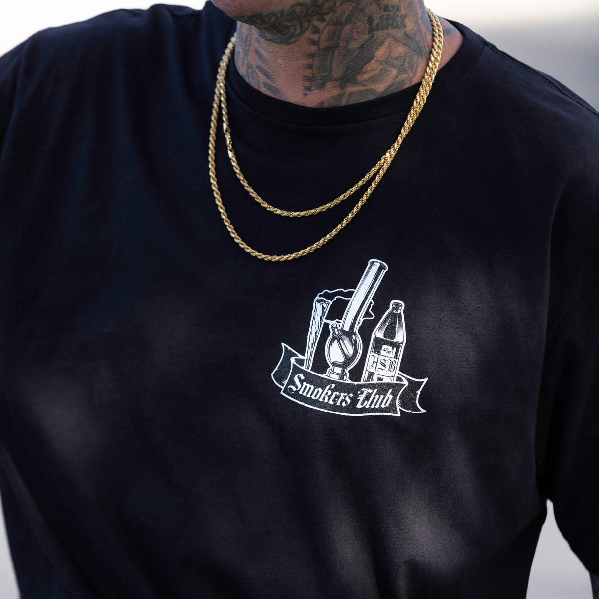Black T-shirt close-up of Smokes Club bottle graphic under a gold chain