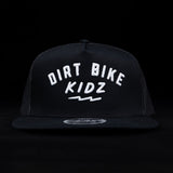 Black trucker cap with white 'DIRT BIKE KIDZ' front graphic and flat brim