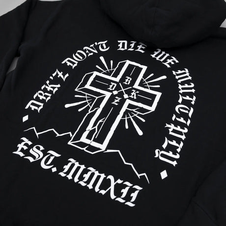 Black hoodie back print: white cross emblem with circular text and rays.
