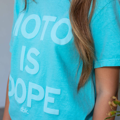 Women’s graphic T-shirt in teal with partial white “Moto is Dope” text print, short sleeves.