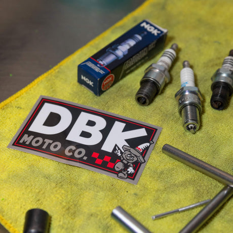 DBK Moto Co sticker with DBK letters and cartoon character, lying on yellow cloth