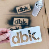 T-shirt stencil card with dbk. logo cutout in hand, with spray paint nearby