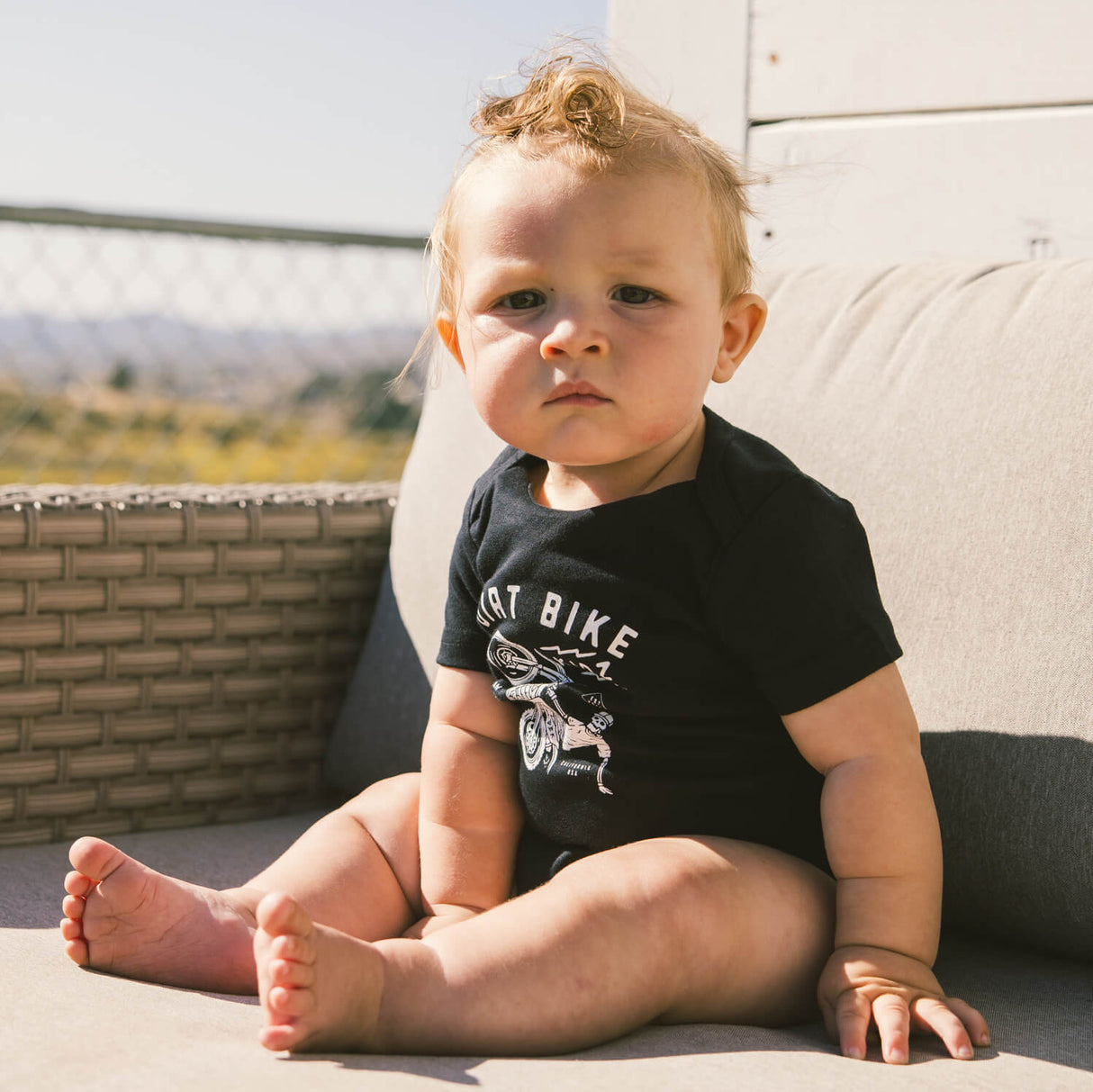 Black baby onesie worn by seated baby, short sleeves, bike graphic on chest