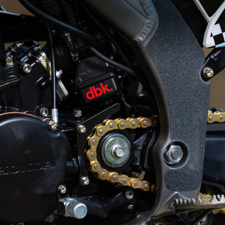 “dbk” logo on dark fabric with gold chain and engine-style artwork