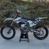 Gray and black motocross dirt bike with knobby tires, gold forks, and stand.
